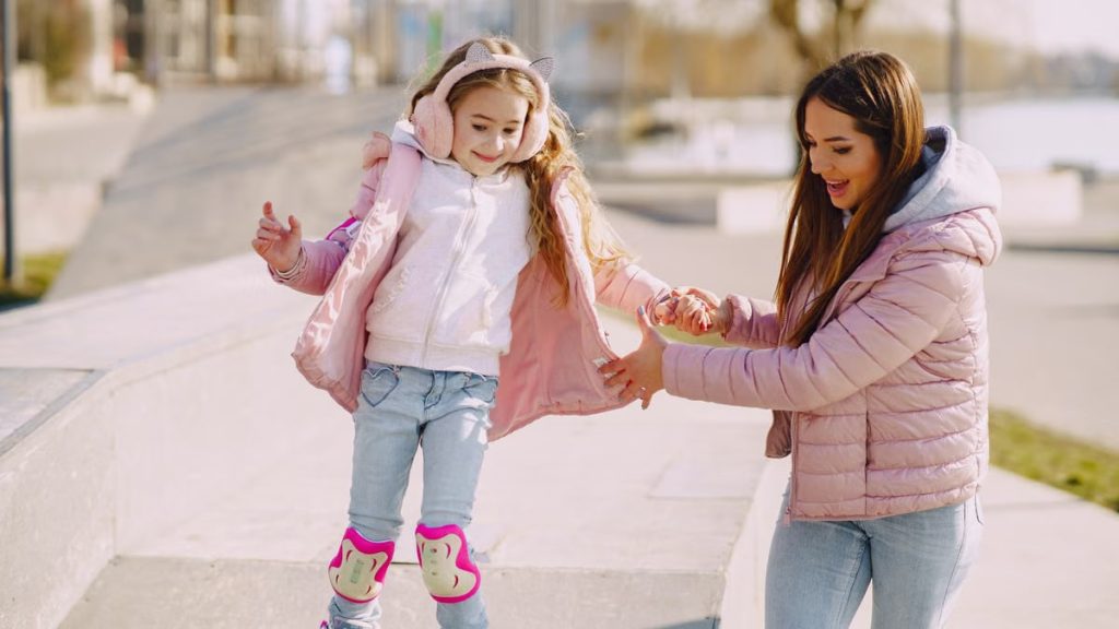 Excited young woman preventing from falling down daughter wearing protection pads while roller skating on concrete ramp during active leisure in sunny spring day