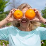 A smiling woman playfully holding oranges in a sunny park setting.