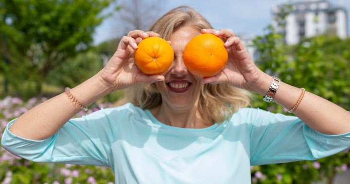 A smiling woman playfully holding oranges in a sunny park setting.